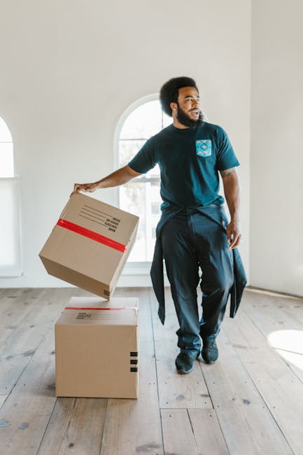 A man with dark curly hair and a beard, dressed in a navy blue uniform with a logo on his chest, is inside a bright, empty room with white walls and large arched windows. He is holding a cardboard box