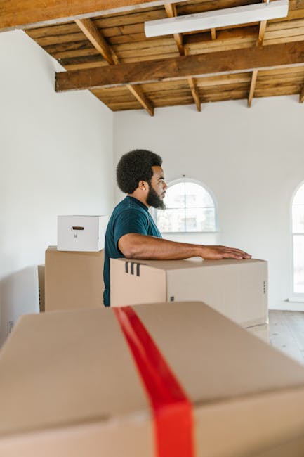 A man with dark curly hair and a beard, dressed in a navy blue uniform with a logo on his chest, is inside a bright, empty room with white walls and large arched windows. He is holding a cardboard box