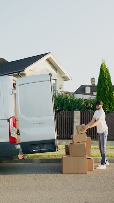 A man wearing a yellow and gray jacket, black cap, and face mask is opening the side door of a white cargo van parked on a brick driveway in a residential area. The van belongs to Man with Van Harold 
