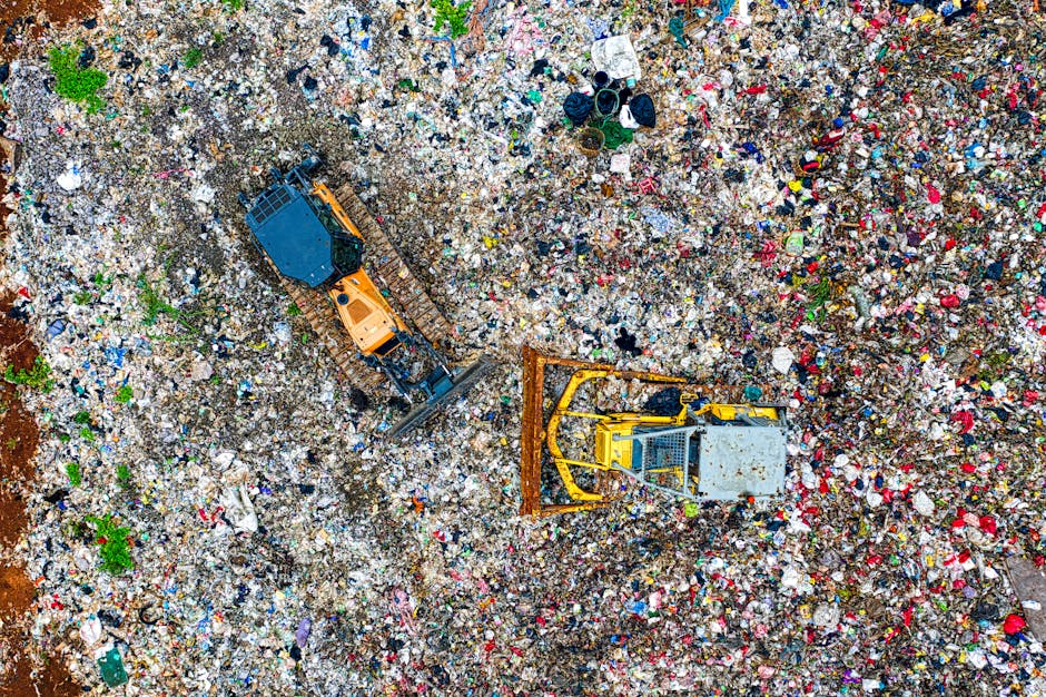 A large landfill site filled with various types of waste materials, including plastic bottles, paper, metal scraps, and other debris, forming multiple mounds across the area. In the background, there is a line of trees and distant high-rise buildings under a cloudy sky. The image emphasizes the scale of waste accumulation, with the foreground showing densely packed refuse and the horizon featuring the urban skyline. The scene is captured from an elevated perspective, highlighting the extent of rubbish disposal and environmental impact. Although the image does not directly depict removals or packing activities, it relates to waste management and disposal processes associated with house relocations and moving services offered by Man with Van Harold Hill, especially when dealing with bulky or unwanted items during a move or clean-out.