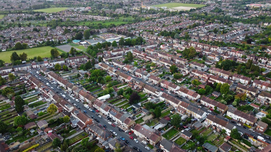 An aerial view of a residential neighborhood showing multiple rows of terraced houses with small front gardens, parked cars lining the streets, and a variety of trees and green spaces interspersed between the housing. In the foreground, a man from Man with Van Harold Hill is loading a large cardboard box wrapped in plastic onto a trolley on the pavement, preparing for a home relocation or furniture transport. Behind him, other boxes and household items are arranged, indicating an ongoing packing and moving process within the property. The background includes more houses, greenery, and open fields, with soft natural lighting highlighting the orderly and tidy area, reflecting professional removals and the logistical planning involved in local house removals within Havering.