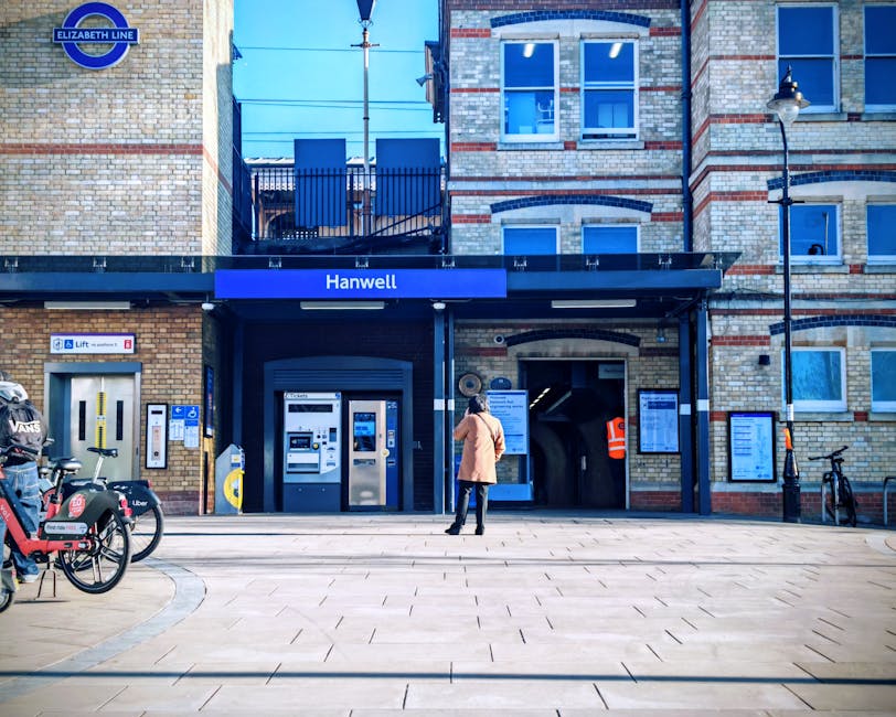 The image shows the entrance to Hanwell Underground station, with a person standing on the pavement facing the station's ticket machines and entrance archway. The station building is constructed from light-colored brick with dark blue trim around the doorway and windows. A blue sign displaying the station name 'Hanwell' is mounted above the entrance, along with the Elizabeth Line logo in the top left corner. To the left of the entrance, there are several bicycles parked, including one with a waterproof cover and a Metro-style rental bike. The area is illuminated by a street lamp on the right side of the image. Inside the station's entrance, the ticket machines are visible, and the station's automated systems can be seen. The overall scene is well-lit with clear skies, indicating daytime. This setting could relate to home relocation or transport logistics managed by [COMPANY_NAME], involved in packing, handling, and loading processes typical of removals and moving services, especially within the context of London transportation infrastructure.