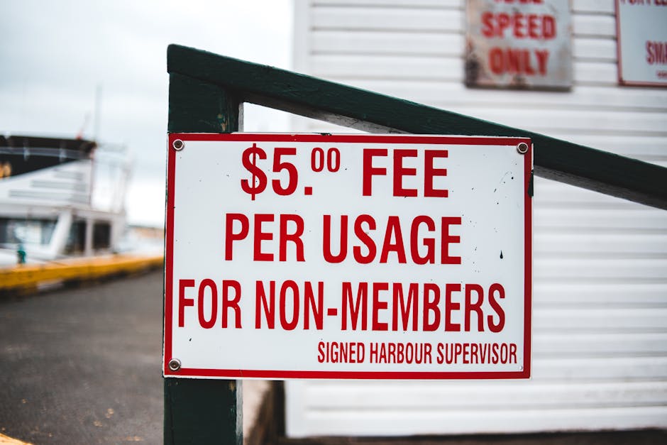 Close-up of a rectangular white sign with bold red text displaying a fee of $5.00 per usage for non-members, attached to a green metal railing near a harbour or marina area. In the background, a section of a boat and a yellow barrier are visible on a paved surface. The sign is positioned outside a building with siding, and there are additional signs partially visible behind it, indicating speed restrictions or other rules. The environment appears overcast, with natural lighting highlighting the sign's details, which relate to harbour regulations and access fees, relevant to transportation and moving logistics handled by Man with Van Harold Hill.