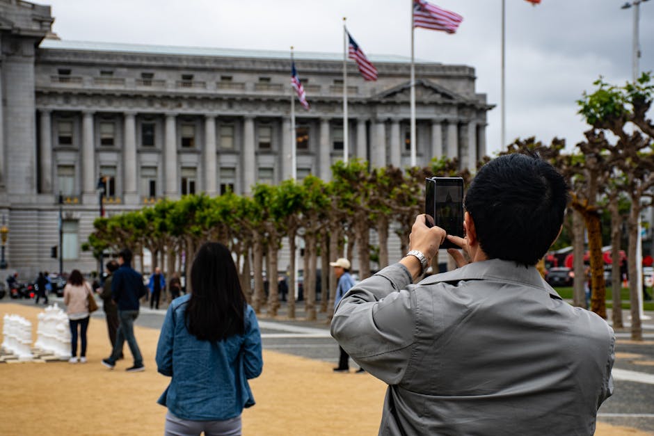 A man with black hair, wearing a grey jacket, is standing outdoors taking a photo with a smartphone in front of a government building with tall columns and American flags waving above. In the foreground, a woman with long dark hair, dressed in a blue jacket and grey pants, faces away from the camera, observing the scene. Several other people are walking around on the paved area, which features neatly trimmed trees without leaves, possibly pruned for aesthetic purposes. The scene occurs during daytime with overcast skies, and the location appears to be a public area or park near the building. The setting suggests a busy urban environment with movement, and the architectural background indicates a significant or historic structure, relevant to location-based content on house removals or relocation services offered by Man with Van Harold Hill.