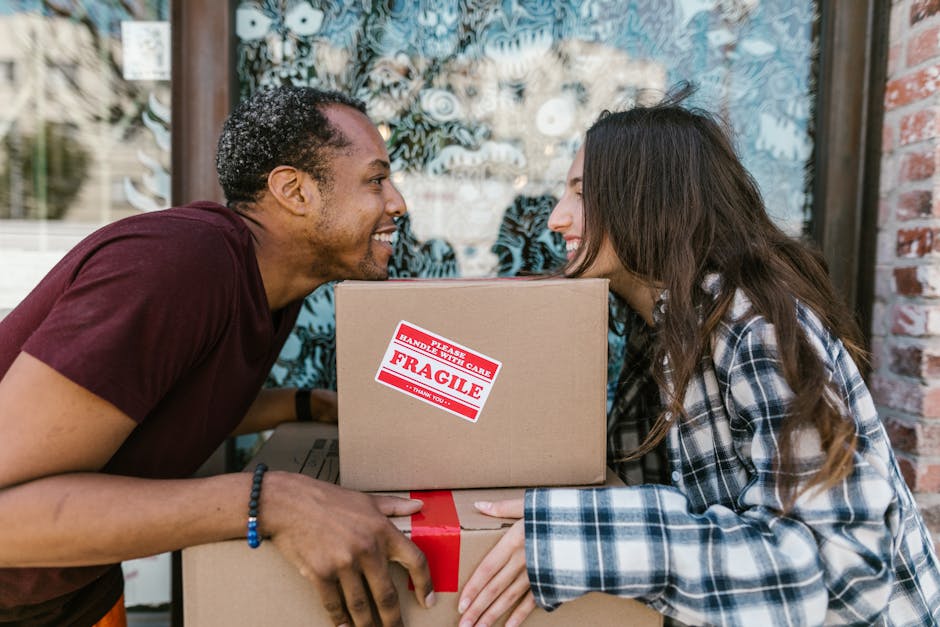 A man and a woman are holding several cardboard moving boxes outdoors, close to a brick building with a large window. The man, on the left, is wearing a dark red T-shirt and a blue beaded bracelet, an