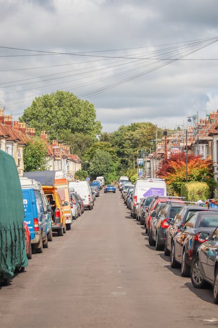 A narrow residential street in Harold Hill with parked cars lining both sides of the roadway, including various small vans and delivery vehicles used for moving purposes. The street is flanked by terr