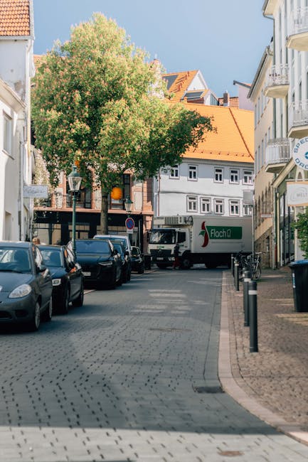 A narrow residential street in Harold Hill with parked cars lining both sides of the roadway, including various small vans and delivery vehicles used for moving purposes. The street is flanked by terr
