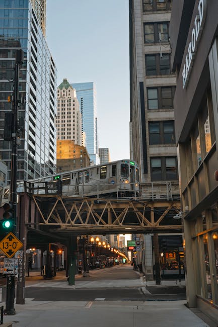A modern urban scene showing an elevated train crossing a busy city street in daylight. The train is metallic with windows and doors visible, moving along a railway bridge supported by a lattice steel structure. Surrounding the scene are tall skyscrapers with glass and stone facades, reflecting the sky and nearby buildings. At street level, traffic lights, a pedestrian crossing signal, and a yellow road sign indicating a height restriction are visible. The sidewalk is clear, with a few street lamps and bus stops, and the overall environment suggests a central business district. The image captures the typical elements of city logistics and transportation, relevant to house removals and furniture transport services provided by companies like Man with Van Harold Hill, highlighting the busy urban setting in which home relocation and moving logistics take place.