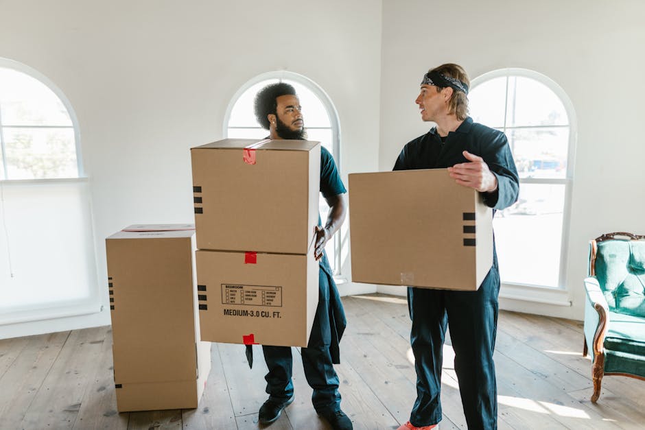 Two movers from Man with Van Harold Hill are inside a room with cream-colored walls and large arched windows, preparing for a home relocation. One mover, with curly hair and wearing a dark T-shirt, is holding a medium-sized cardboard box and is engaged in conversation with the other mover, who has blonde hair, wears a headband, and is dressed in a dark hoodie and pants. The second mover is gesturing with his hand while holding an empty cardboard box. Several other cardboard boxes, of varying sizes, are stacked on the wooden floor, some sealed with red tape, indicating packing and moving preparations. The room is well-lit with natural light coming through the windows, and a vintage-style green armchair with wooden accents is located to the right. The furniture, packing materials, and the presence of manual handling suggest the loading process of a furniture transport and household move, typical of local removal services provided by [COMPANY_NAME].