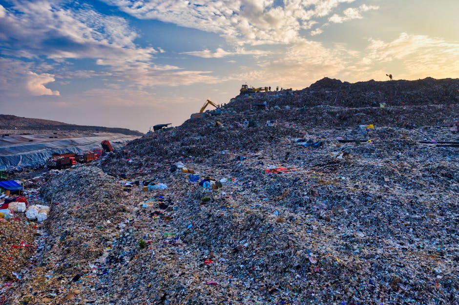 A large landfill site filled with heaps of mixed waste materials including plastic, paper, metal, and textiles, extending across the landscape under a partly cloudy sky during late afternoon or early evening. At the top of the mound, construction equipment such as an excavator is visible, indicating ongoing waste management operations. The foreground shows smaller piles of refuse, with discarded packaging, plastic containers, and other debris scattered throughout the area. In the background, there are patches of tarpaulin covers and cranes, suggesting active sorting or loading processes. The scene is illuminated with natural lighting, creating a contrast between the dark, piled-up waste and the brighter sky, highlighting the scale of waste accumulation. This environmental image reflects waste disposal and recycling logistics often involved in property clearance or site cleanups that Man with Van Harold Hill may coordinate as part of comprehensive removals services.