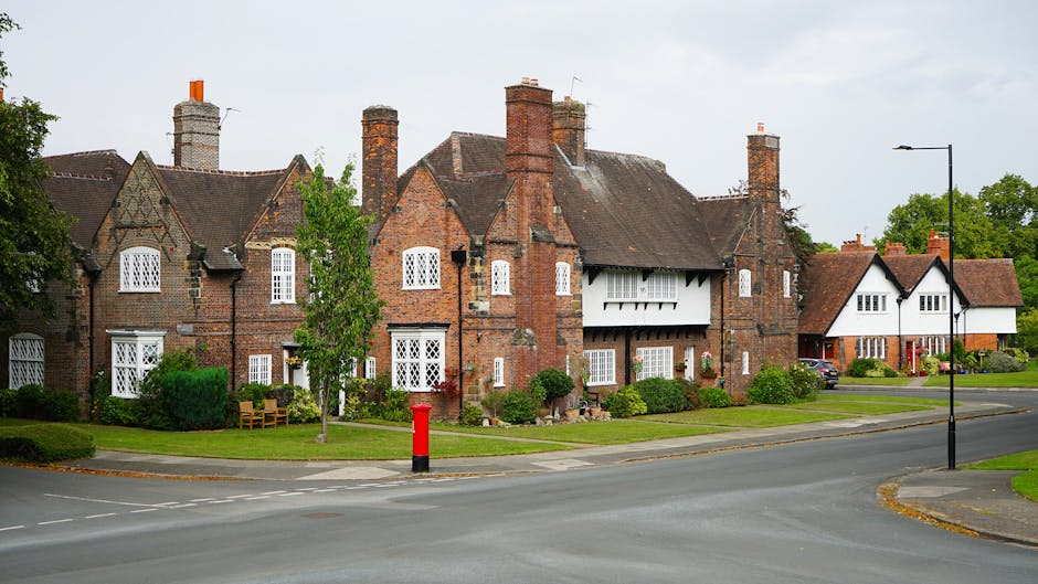 A row of traditional brick houses with pitched roofs and multiple chimneys, located along a residential street. The properties feature white-framed windows, some with decorative lattice designs, and small front gardens with neatly trimmed bushes and trees. One house has a white balcony under its chimney, and another displays a white gabled frontage. The street is paved, with a red post box situated near the curb, and a black street lamp on the sidewalk. The area appears calm and well-maintained, suitable for home relocation and furniture transport activities. The image showcases an outdoor setting during daylight hours, with some greenery in the background, reflecting a typical residential environment that [COMPANY_NAME], such as Man with Van Harold Hill, might work within for local removals and house moving services.