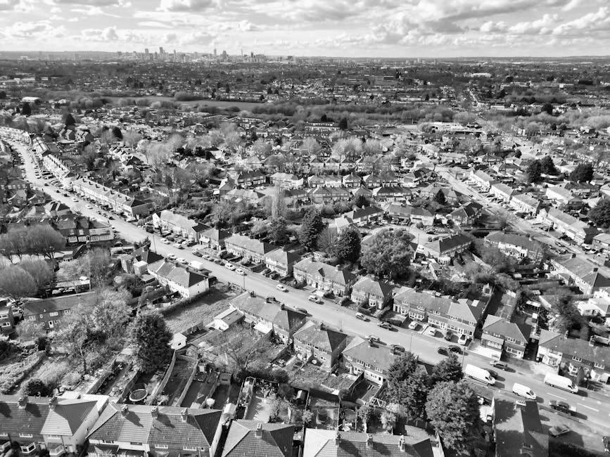 Aerial black-and-white photograph showing a residential neighbourhood with rows of terraced and semi-detached houses, each with small gardens and driveways. A main road runs diagonally through the scene, with parked cars lining both sides. Behind the houses, an expanse of open land or fields extends toward the horizon, where a distant city skyline with tall buildings and structures is visible under a sky filled with clouds. The scene captures a typical suburban area during daylight hours, with no visible moving equipment or vehicles related to removals or furniture transport in the image, focusing solely on the urban landscape and housing arrangements.